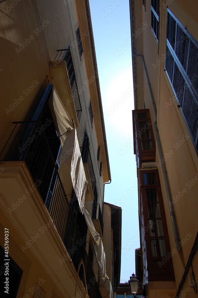 Fototapeta premium Close up outdoor view from above of the top of buildings in a narrow street. Blue sky visible between dark grey walls. Houses facades with windows and shutters.