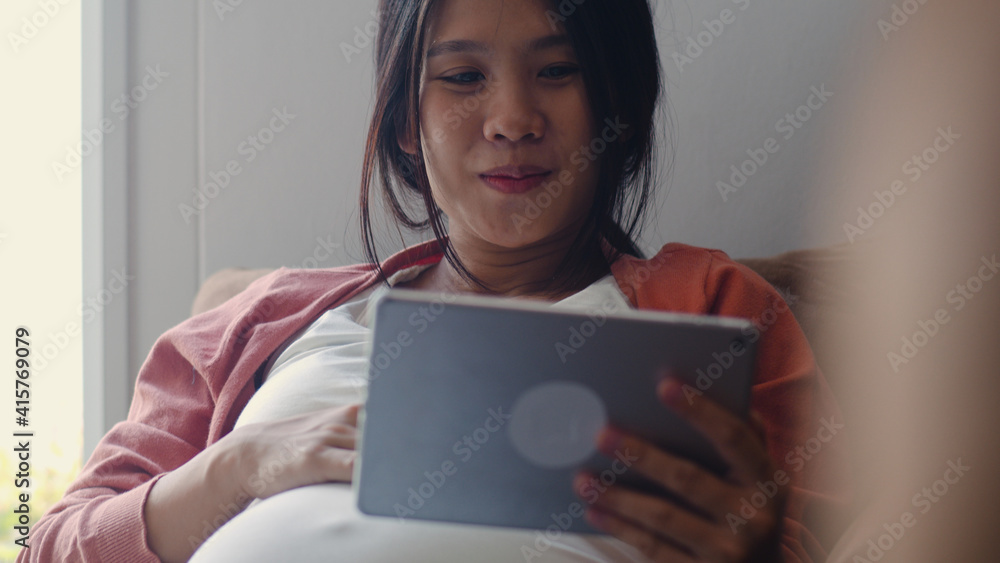 Young Asian Pregnant woman using tablet search pregnancy information. Mom feeling happy smiling positive and peaceful while take care her child lying on sofa in living room at home concept. Young Asian Pregnant woman using tablet search pregnancy information. Mom feeling happy smiling positive and peaceful while take care her child lying on sofa in living room at home concept.