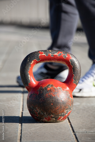 Fototapeta Naklejka Na Ścianę i Meble -  Close up shot of an old, very used red gym iron ball on a paved flloor. Outdoor training session due to covid restrictions and gym closures in Italy. Trainer's feet in the background.