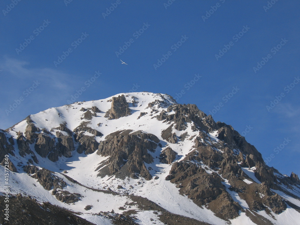Snow covered mountain in the Hautes Alpes region of France, with lone glider flying above the peak on a sunny day in early Spring