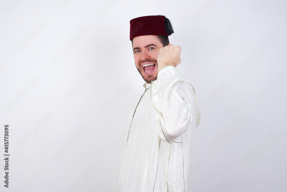 Overjoyed young handsome Caucasian man wearing Arab djellaba and Fez hat over white wall glad to receive good news, clenching fist and making winning gesture.