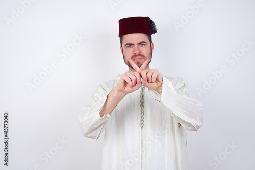 young handsome Caucasian man wearing Arab djellaba and Fez hat over white wall has rejection angry expression crossing fingers doing negative sign.