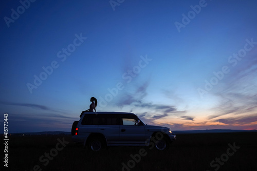 Silhouette of girl with ponytail on roof of car on background of sunset sky. Blue and orange sky. White off road car