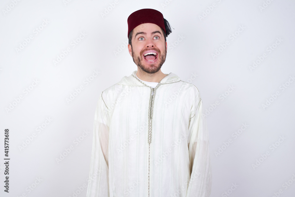 Surprised young handsome Caucasian man wearing Arab djellaba and Fez hat over white wall, shrugs shoulders, looking sideways, being happy and excited. Sudden reactions concept.