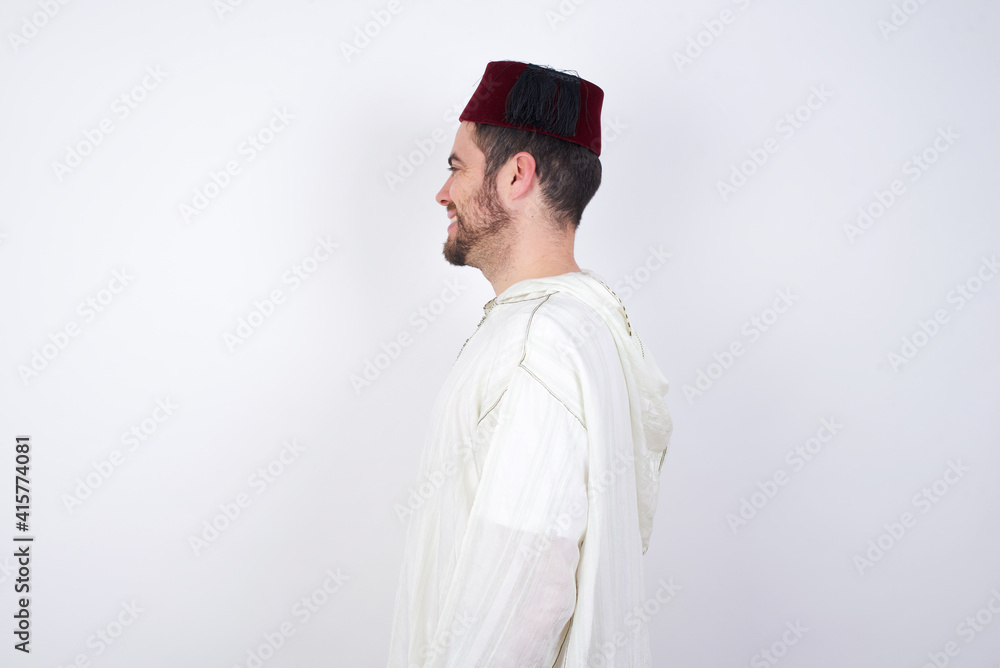 Profile of smiling young handsome Caucasian man wearing Arab djellaba and Fez hat over white wall with healthy skin, has contemplative expression, ready to have outdoor walk. Profile of smiling young handsome Caucasian man wearing Arab djellaba and Fez hat over white wall with healthy skin, has contemplative expression, ready to have outdoor walk.