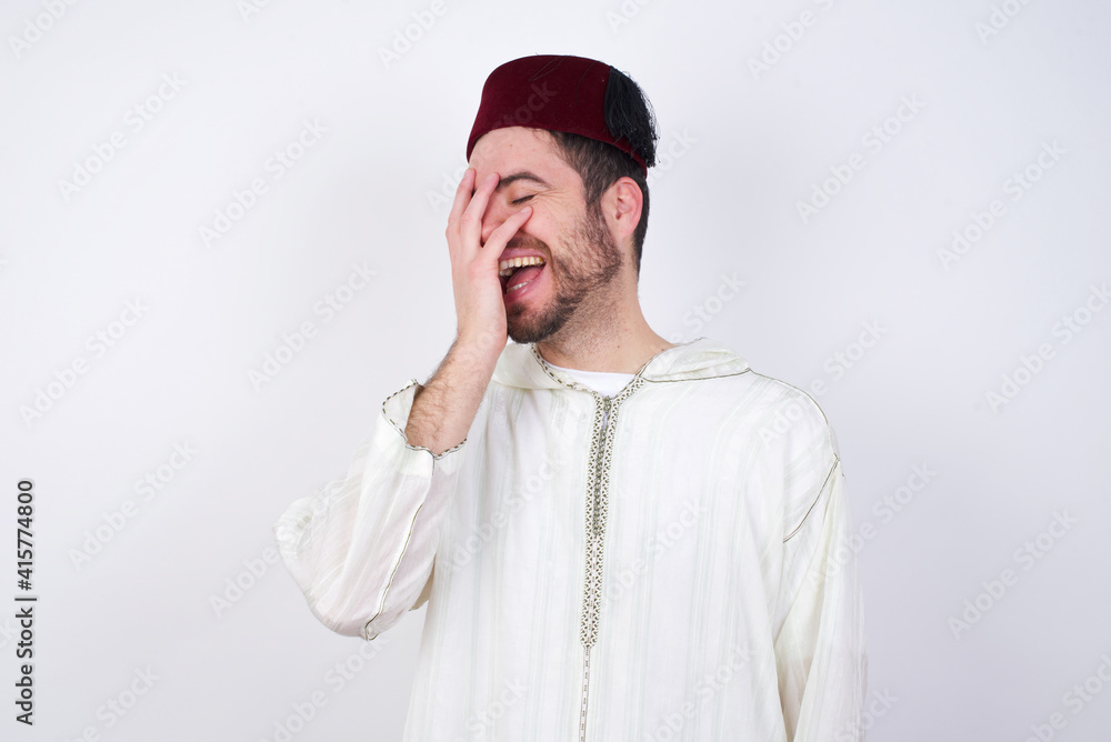 Charismatic carefree joyful young handsome Caucasian man wearing Arab djellaba and Fez hat over white likes laugh out loud not hiding emotions giggling hear funny hilarious joke chuckling facepalm. Charismatic carefree joyful young handsome Caucasian man wearing Arab djellaba and Fez hat over white likes laugh out loud not hiding emotions giggling hear funny hilarious joke chuckling facepalm.