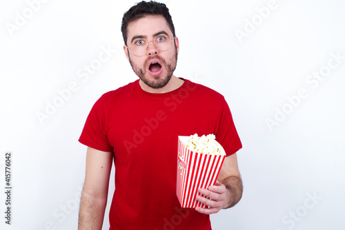 young handsome man in red T-shirt against white background eating popcorn having stunned and shocked look, with mouth open and jaw dropped exclaiming: Wow, I can't believe this. Surprise and shock