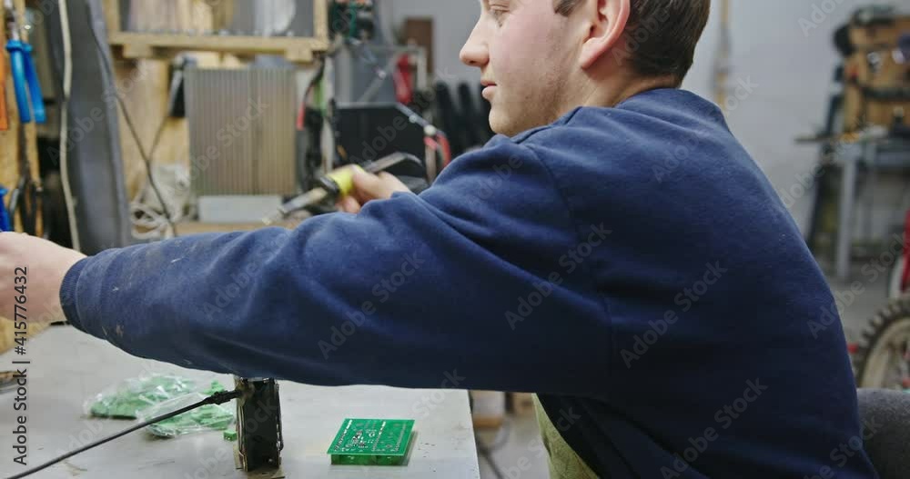 Man soldering microchip in workshop. Side view male technician sitting at table and soldering microchip during work in grungy workshop Man soldering microchip in workshop. Side view male technician sitting at table and soldering microchip during work in grungy workshop