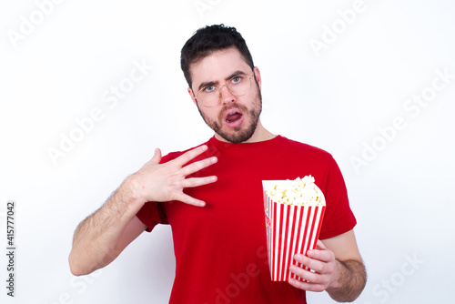young handsome man in red T-shirt against white wall keeps hands on chest feeling shocked and scared, mouth widely opened, stares at camera saying: Who, me?