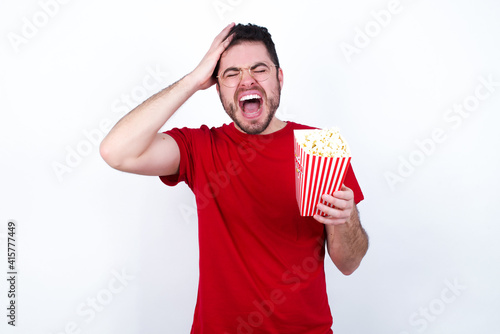 Shocked panic young handsome man in red T-shirt against white background eating popcorn holding hands on head and screaming in despair and frustration.