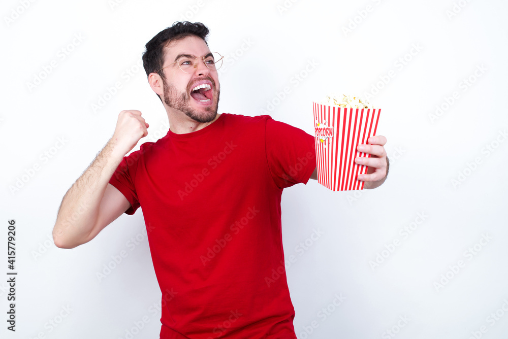 Attractive young handsome man in red T-shirt against white background eating popcorn celebrating a victory punching the air with his fists and a beaming toothy smile Attractive young handsome man in red T-shirt against white background eating popcorn celebrating a victory punching the air with his fists and a beaming toothy smile