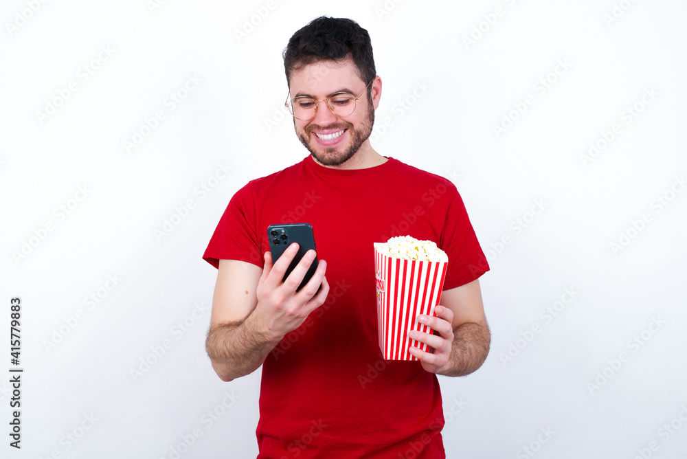 Smiling Young handsome man in red T-shirt against white background eating p using cell phone, messaging, being happy to text with friends, looking at smartphone. Modern technologies and communication. Smiling Young handsome man in red T-shirt against white background eating p using cell phone, messaging, being happy to text with friends, looking at smartphone. Modern technologies and communication.