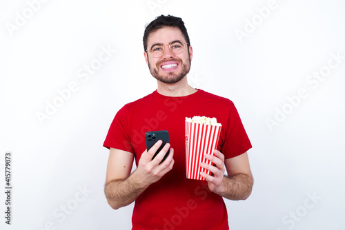 Smiling young handsome man in red T-shirt against white background eating popcorn friendly and happily holding mobile phone taking selfie in mirror.