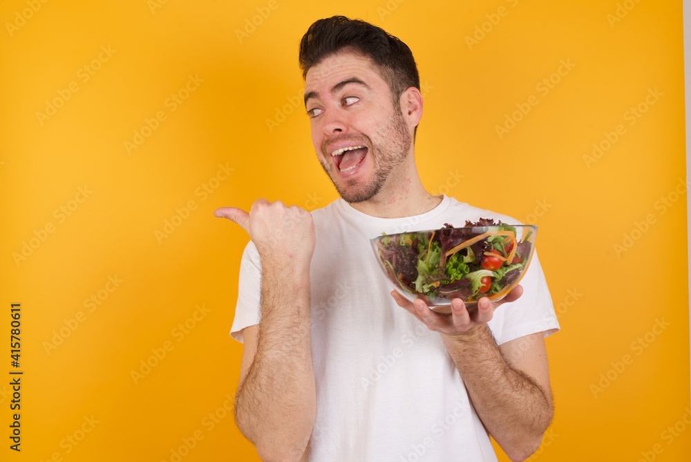 Stupefied young handsome Caucasian man holding a salad bowl against yel with surprised expression, opens eyes and mouth widely, points aside with thumb, shows something strange. Advertisement concept. Stupefied young handsome Caucasian man holding a salad bowl against yel with surprised expression, opens eyes and mouth widely, points aside with thumb, shows something strange. Advertisement concept.