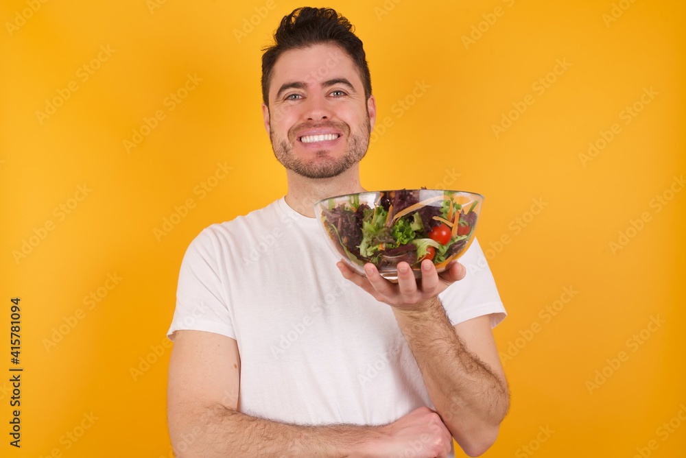 Cheerful young handsome Caucasian man holding a salad bowl against yellow wall with hand near face. Looking with glad expression at the camera after listening to good news. Confidence. Cheerful young handsome Caucasian man holding a salad bowl against yellow wall with hand near face. Looking with glad expression at the camera after listening to good news. Confidence.