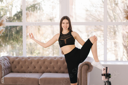 Young athletic woman blogger in balck sportswear shoots video on phone as she does exercises at home in living room. Sport and recreation concept. Fitness vlogger recording live tutorial video © Aleksandr
