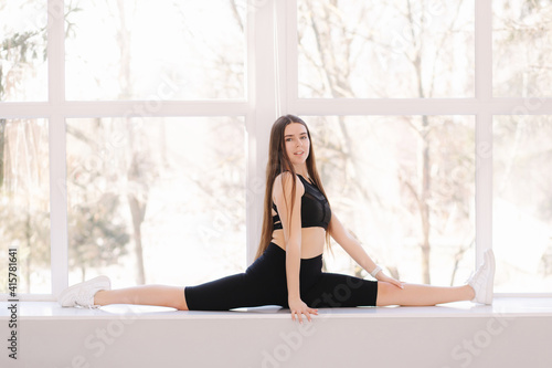 Beautifull brunette woman doing twine stretching on windowsill at home. Attractive fit girl training at home. Copy space