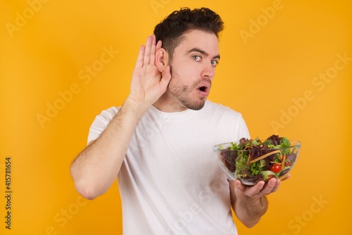 Oh my God! Funny astonished young handsome Caucasian man holding a salad bowl against yellow wall opening mouth widely looking aside, with hand near ear trying to listen to gossips.