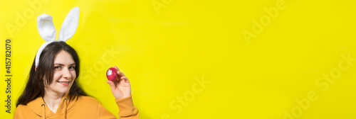 A happy brunette woman with rabbit ears on her head holds Easter eggs and smiles. Prepare for Easter, on a yellow background banner with copy space for text.