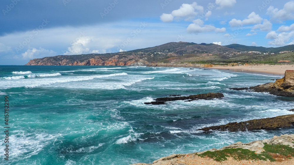 Amazing view to the blue Atlantic ocean, with big waves, and sandy Guincho Beach. Stormy clouds and dark mountain of Sintra-Cascais natural park, visible in the distance. Beautiful landscape. Portugal