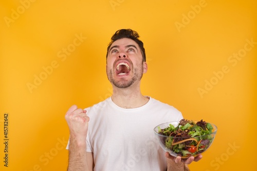 young handsome Caucasian man holding a salad bowl against yellow wall looks with excitement up, keeps hands raised, notices something unexpected.