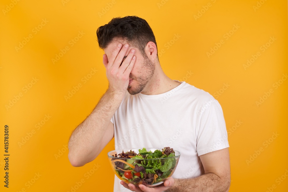 Sad young handsome Caucasian man holding a salad bowl against yellow wall covering face with hands and crying.