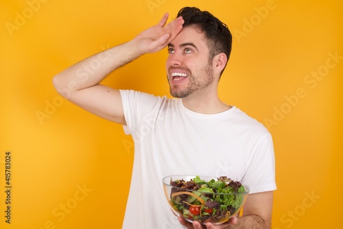 young handsome Caucasian man holding a salad bowl against yellow wall touching forehead  hears something surprising  glad receive good news  feels relieved. Almost got in trouble.