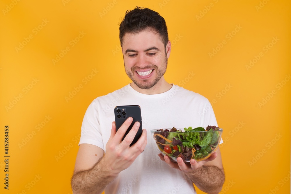 Smiling young handsome Caucasian man holding a salad bowl against yellow using cell phone, messaging, being happy to text with friends, looking at smartphone. Modern technologies and communication. Smiling young handsome Caucasian man holding a salad bowl against yellow using cell phone, messaging, being happy to text with friends, looking at smartphone. Modern technologies and communication.