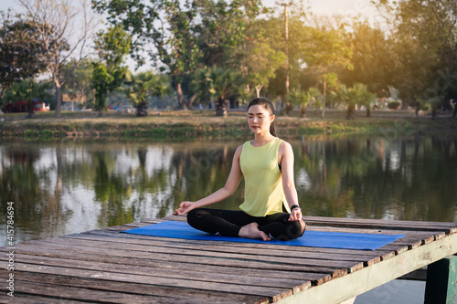 Young Asian beautiful woman practicing yoga and meditate in the lotus pose outdoor beside the lake in the morning for relaxation and peace of mind. Harmony and meditation concept. Healthy lifestyle