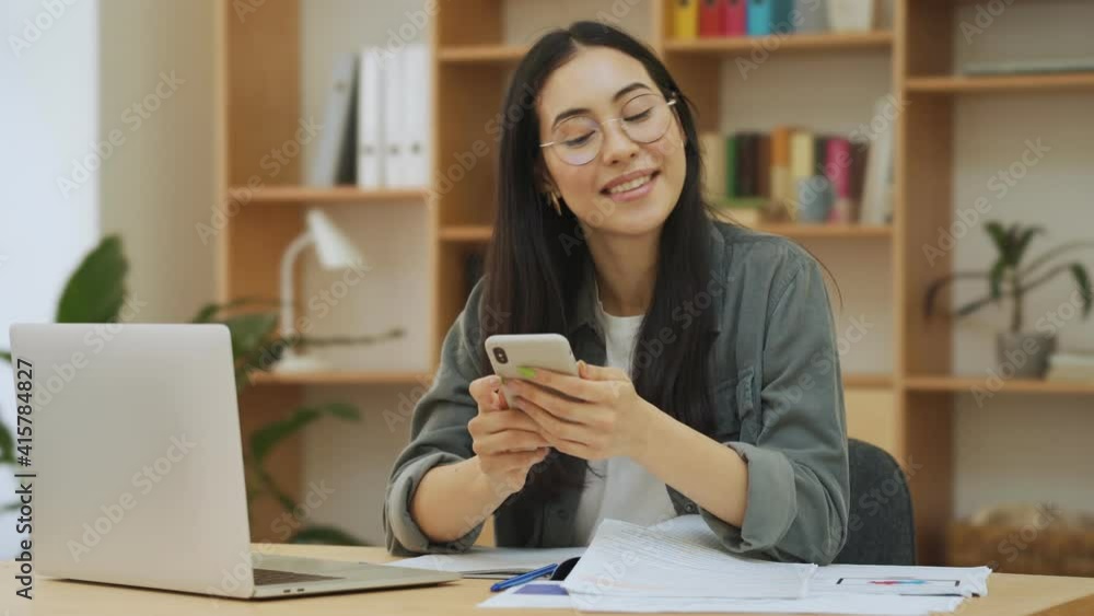 A smiling young asian korean woman is working using her smartphone and laptop computer while sitting in the office A smiling young asian korean woman is working using her smartphone and laptop computer while sitting in the office