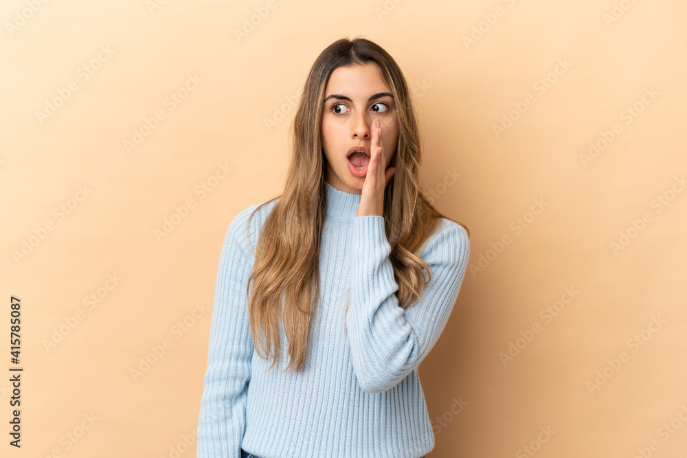 Young caucasian woman isolated on beige background whispering something with surprise gesture while looking to the side Young caucasian woman isolated on beige background whispering something with surprise gesture while looking to the side