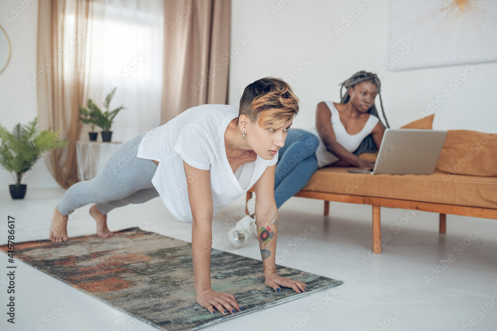 Short-haired young girl doing yoga at home while her girlfriend sitting on the sofa