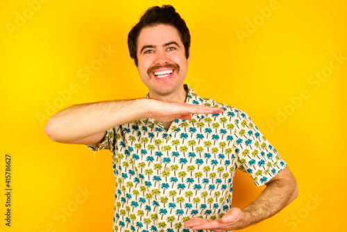 young handsome Caucasian man wearing Hawaiian shirt against yellow wall gesturing with hands showing big and large size sign, measure symbol. photo