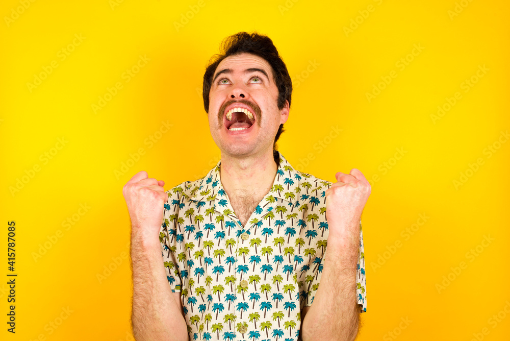 young handsome Caucasian man wearing Hawaiian shirt against yellow wall looks with excitement up, keeps hands raised, notices something unexpected. young handsome Caucasian man wearing Hawaiian shirt against yellow wall looks with excitement up, keeps hands raised, notices something unexpected.
