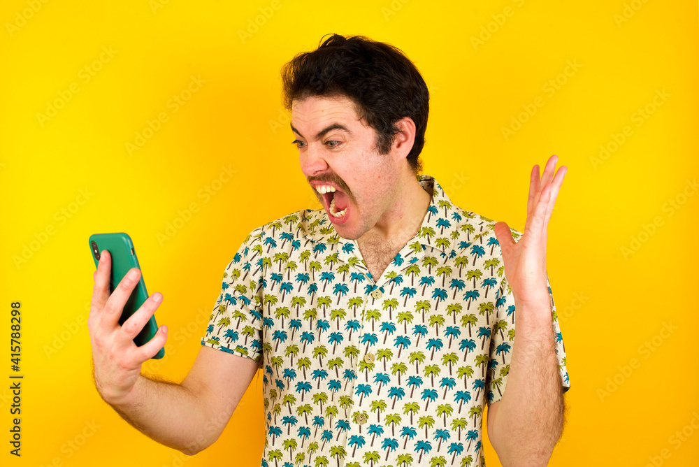Photo of outraged annoyed young handsome Caucasian man wearing Hawaiian shirt against yellow wall holds cell phone, makes call, argues with colleague, expresses negative emotions. People and anger. Photo of outraged annoyed young handsome Caucasian man wearing Hawaiian shirt against yellow wall holds cell phone, makes call, argues with colleague, expresses negative emotions. People and anger.