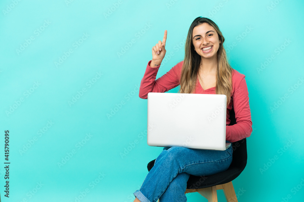 Young caucasian woman sitting on a chair with her pc isolated on blue background showing and lifting a finger in sign of the best