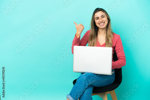 Young caucasian woman sitting on a chair with her pc isolated on blue background pointing to the side to present a product © luismolinero