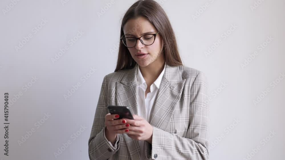 A happy young businesswoman wearing glasses is using a smartphone while doing a winner gesture standing isolated over grey background in the studio A happy young businesswoman wearing glasses is using a smartphone while doing a winner gesture standing isolated over grey background in the studio