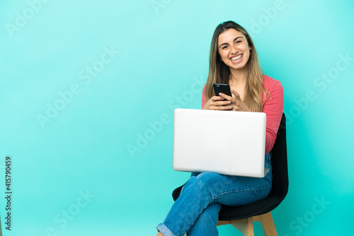 Young caucasian woman sitting on a chair with her pc isolated on blue background sending a message with the mobile © luismolinero