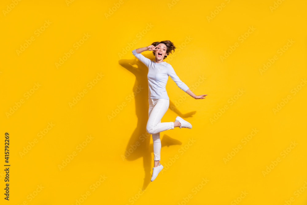 Full length photo of pretty funny young woman wear white shirt jumping showing v-signs cover eye isolated yellow color background