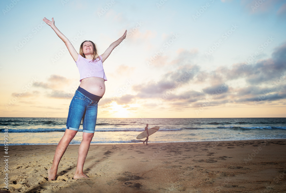 Happy calm pregnant woman during vacations on the sand beach lift hands with surfer holding surfboard pass on the back Happy calm pregnant woman during vacations on the sand beach lift hands with surfer holding surfboard pass on the back