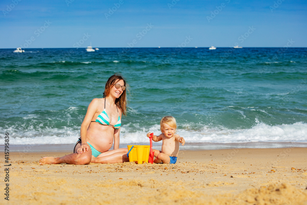 Toddler boy sit and pregnant young mother on the sand beach playing together near sea waves pointing with finger Toddler boy sit and pregnant young mother on the sand beach playing together near sea waves pointing with finger