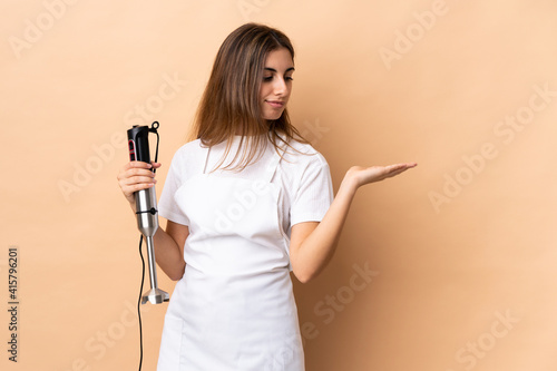 Woman using hand blender over isolated background holding copyspace with doubts