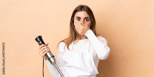 Woman using hand blender over isolated background covering mouth with hand