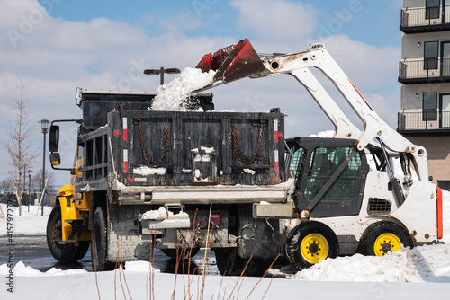 a large metal bucket of a tracked excavator loads snow from a city road onto a truck body