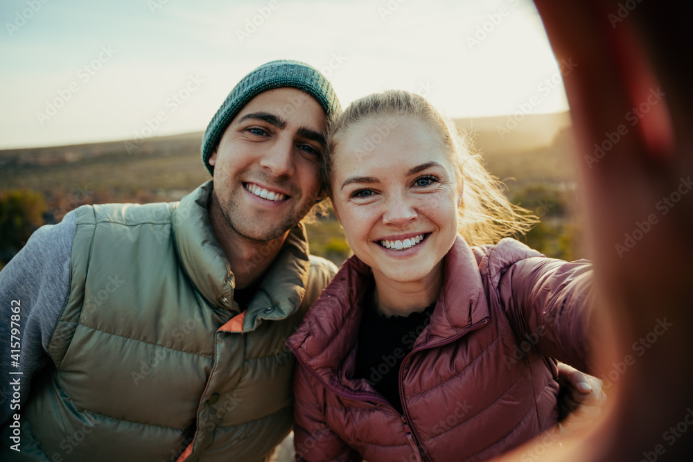 Caucasian husband and wife taking selfie standing on top of mountain smiling while sun sets behind