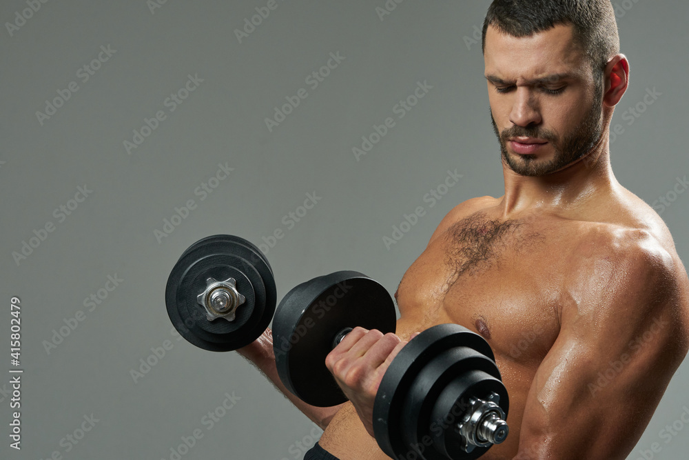 Focused bearded Caucasian sportsman building up muscles with dumbbells in room indoors Focused bearded Caucasian sportsman building up muscles with dumbbells in room indoors