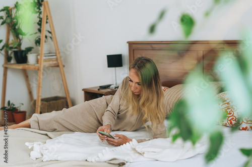 Beautiful blonde young woman using mobile on bed in bright room at home photo