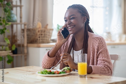 Positive Black Lady Talking On Cellphone And Enjoying Tasty Breakfast In Kitchen