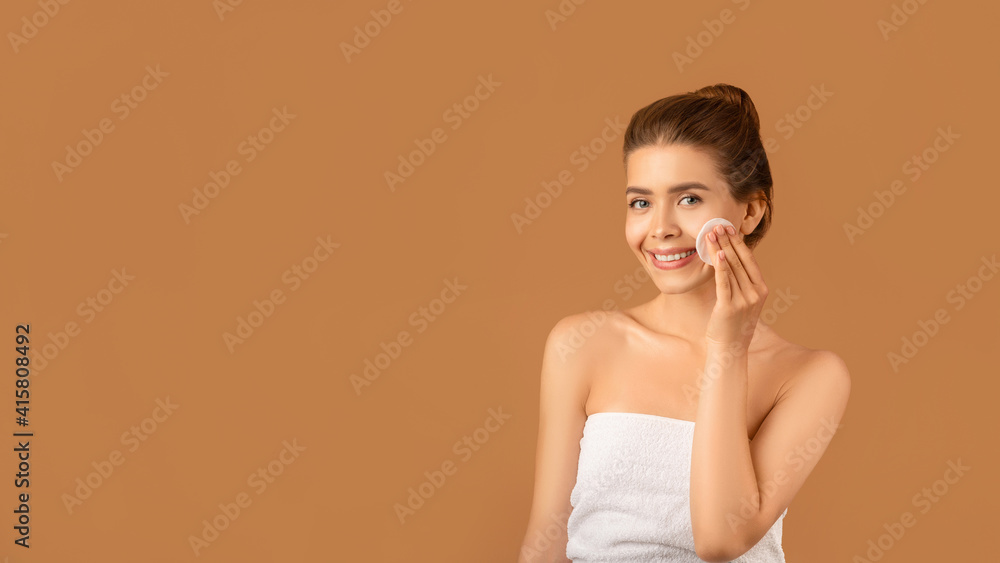 Pretty young lady wiping her face with cotton pad, cleansing her skin with micellar water or tonic on brown background Pretty young lady wiping her face with cotton pad, cleansing her skin with micellar water or tonic on brown background
